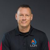 Man in a black shirt with a logo, posing against a dark background.